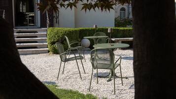 A courtyard with outdoor seating, including round tables and chairs, surrounded by a gravel area and a hedge.