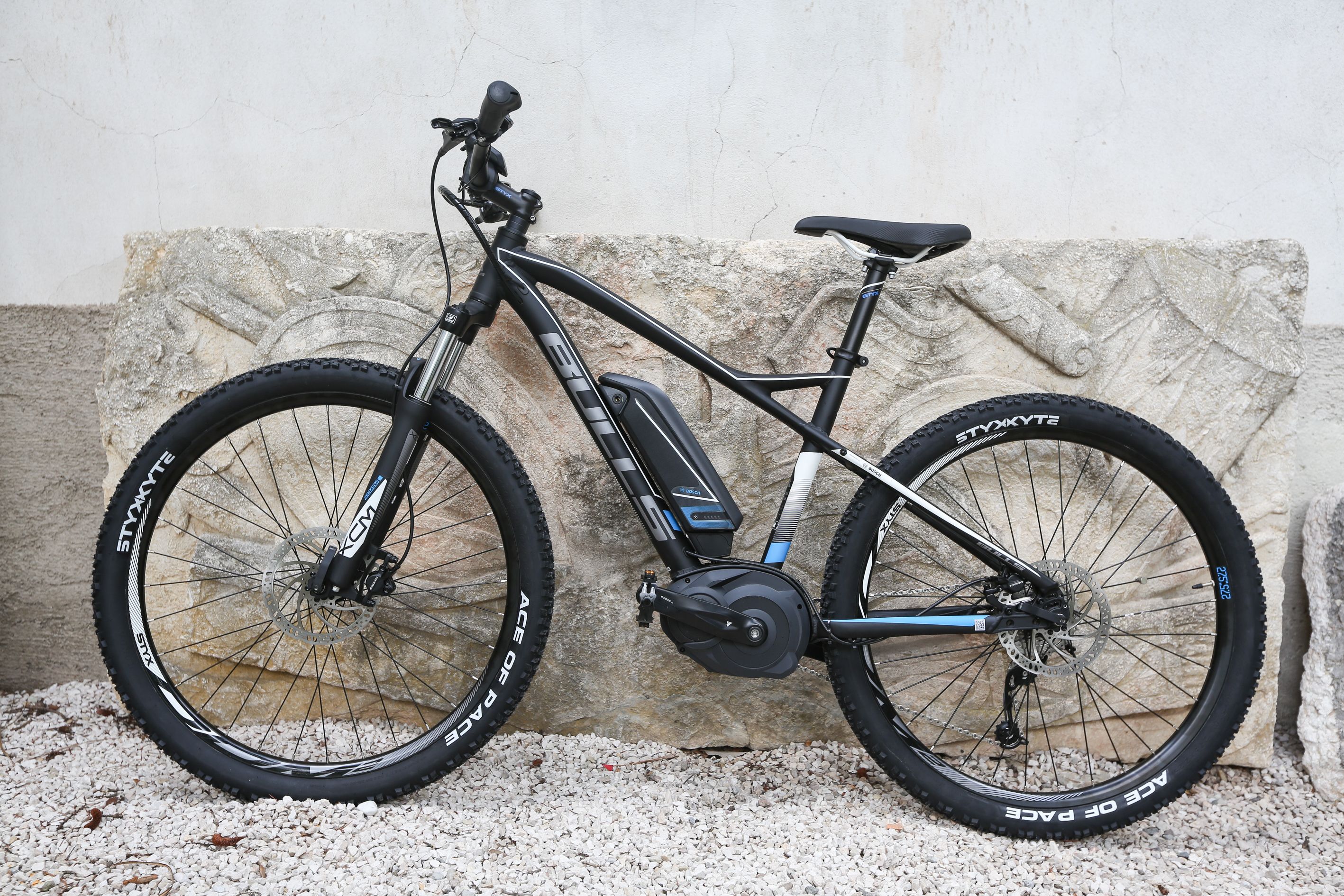 Ein feststehendes E-Bike mit schwarzem Rahmen und blauen Akzenten, das an einer Steinmauer steht.