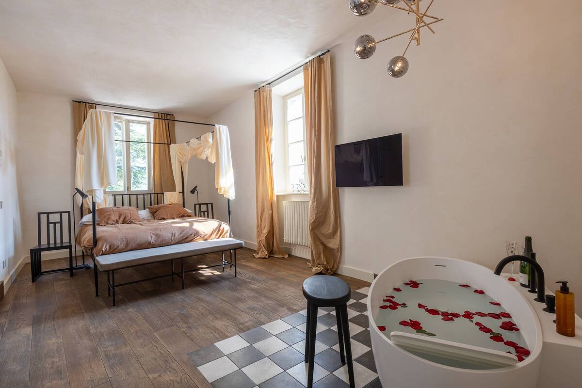 A bedroom with a bed, a bench, a bathtub with rose petals, a TV, and a window with curtains.