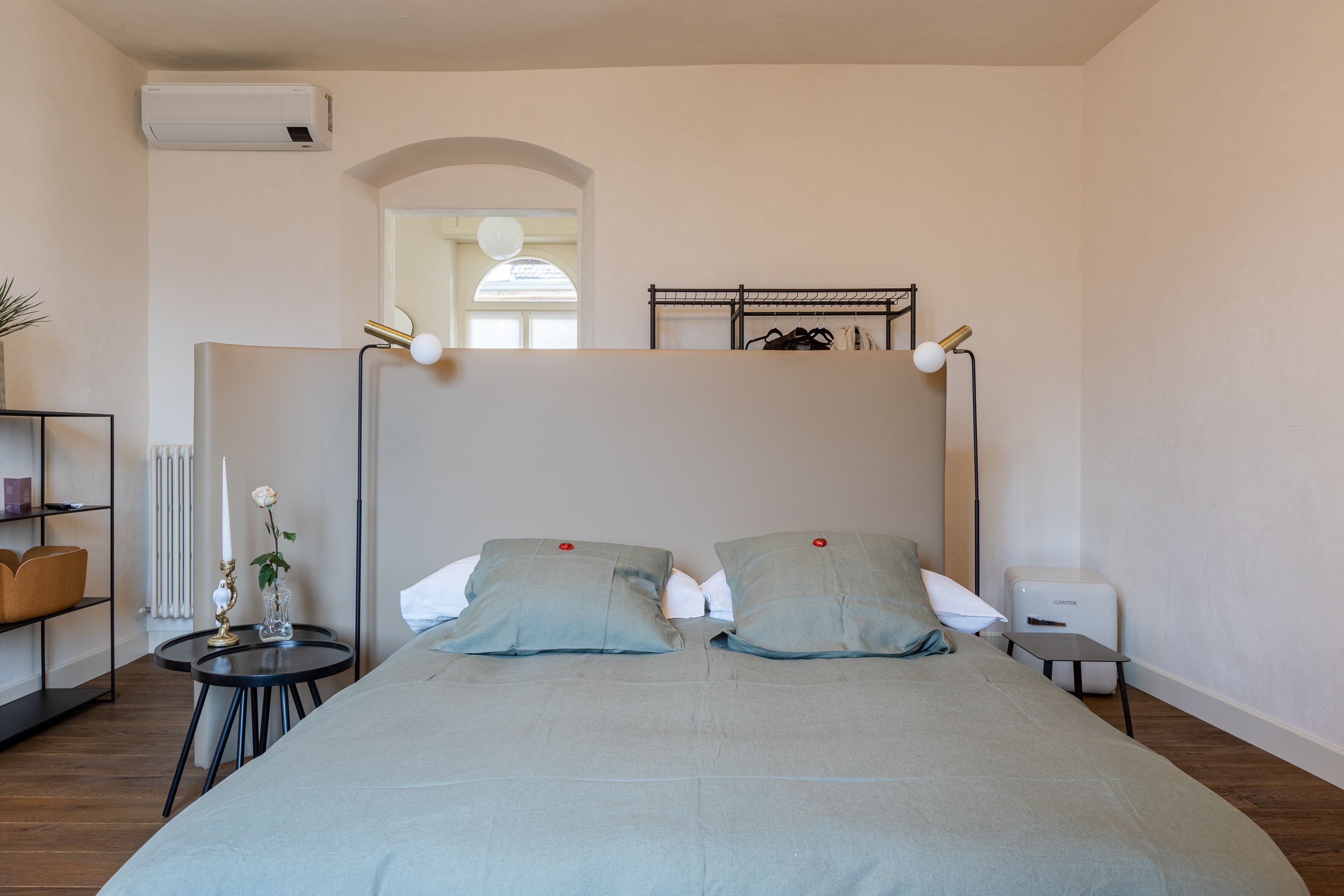 Un dormitorio con una cama, mesitas de noche, un radiador, una estantería con libros y una ventana.