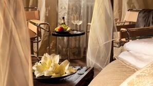 Una habitació d'hotel amb un llit, una taula de nit amb una ampolla i gots, i un safata amb fruites i flors.