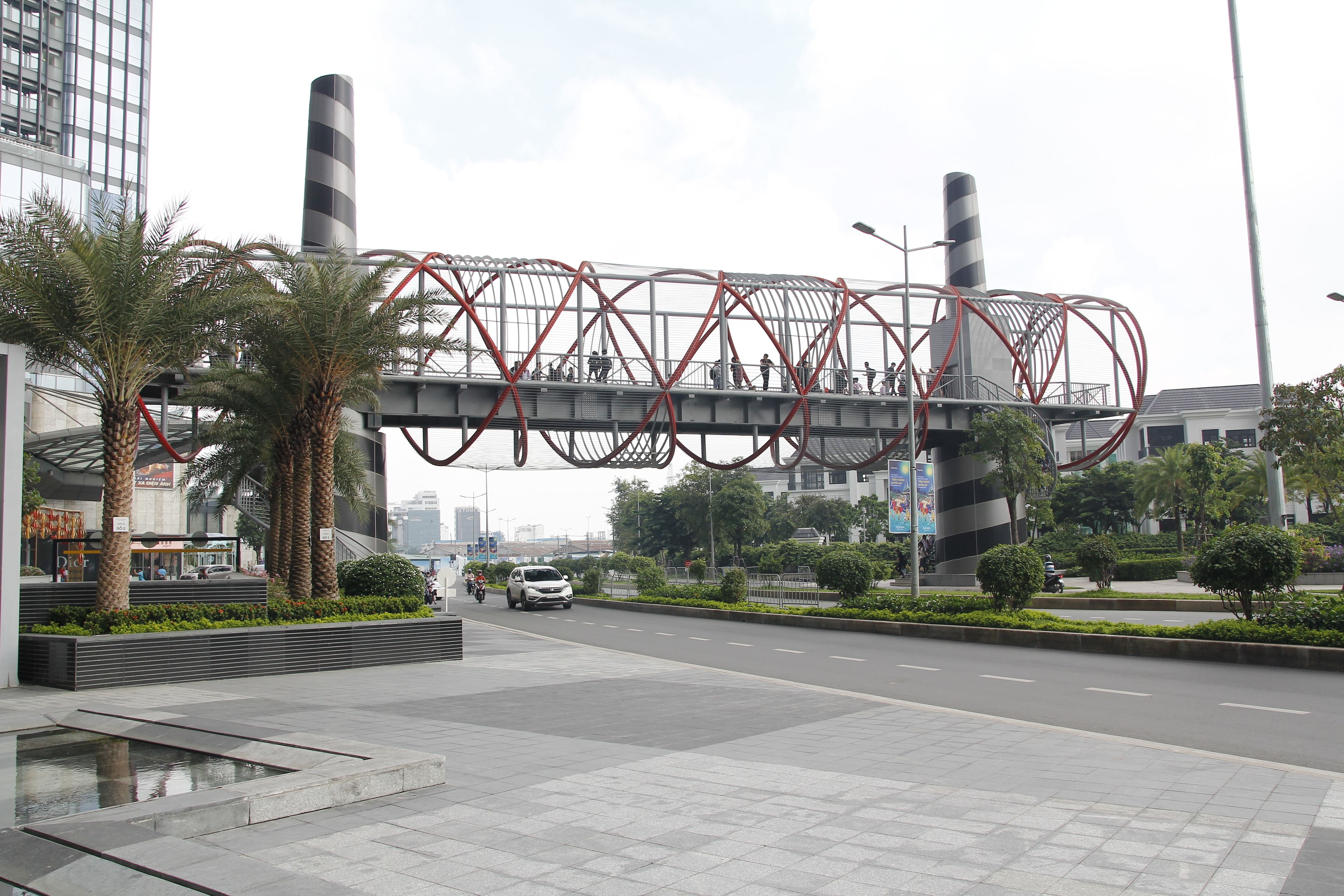 A modern urban street with a pedestrian bridge, palm trees, and a road with vehicles.