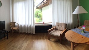 Una habitación con piso de madera, televisor, sofá, mesa y ventana con cortinas.