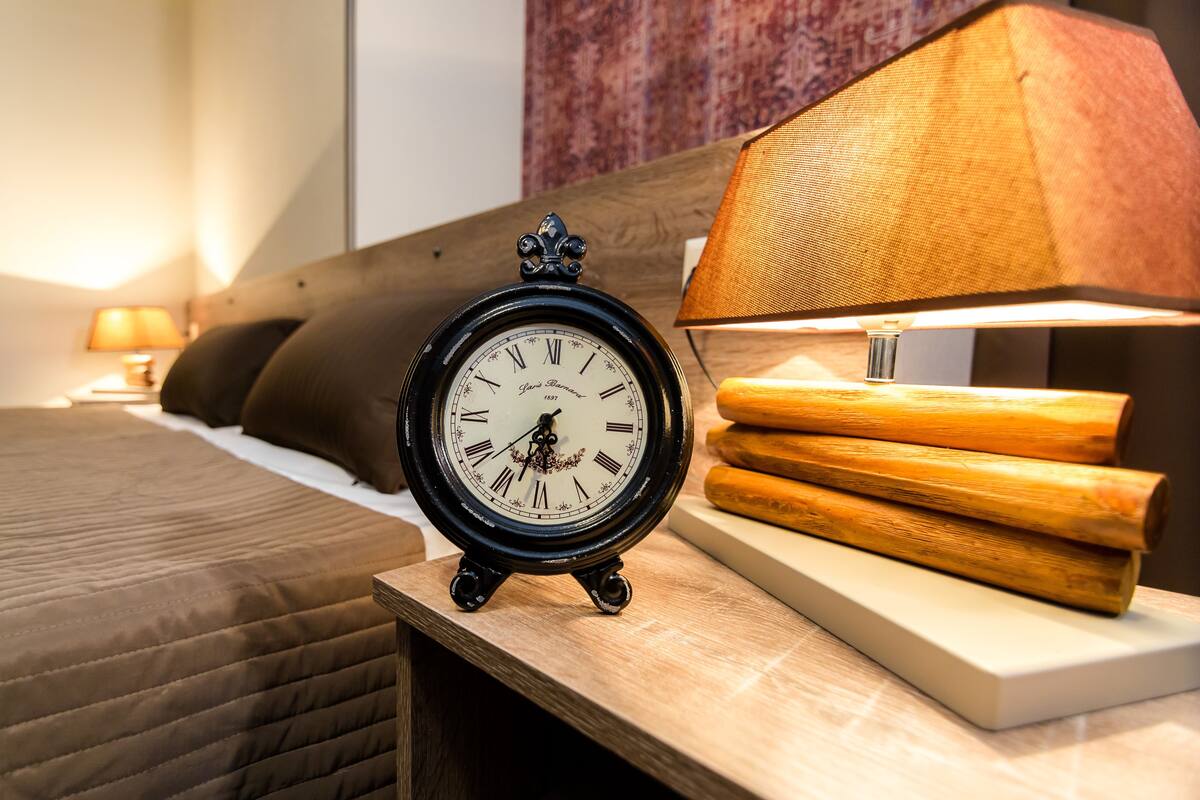 A bed with a quilted cover, a black wall clock, a wooden nightstand, and a lamp with a beige shade.