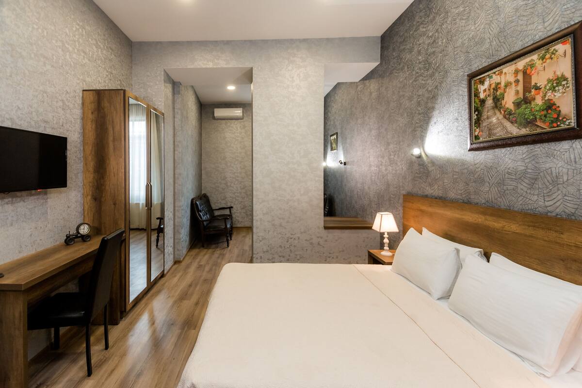 A hotel room with a wooden desk, a bed with white linens, a TV mounted on the wall, and a painting above the bed.