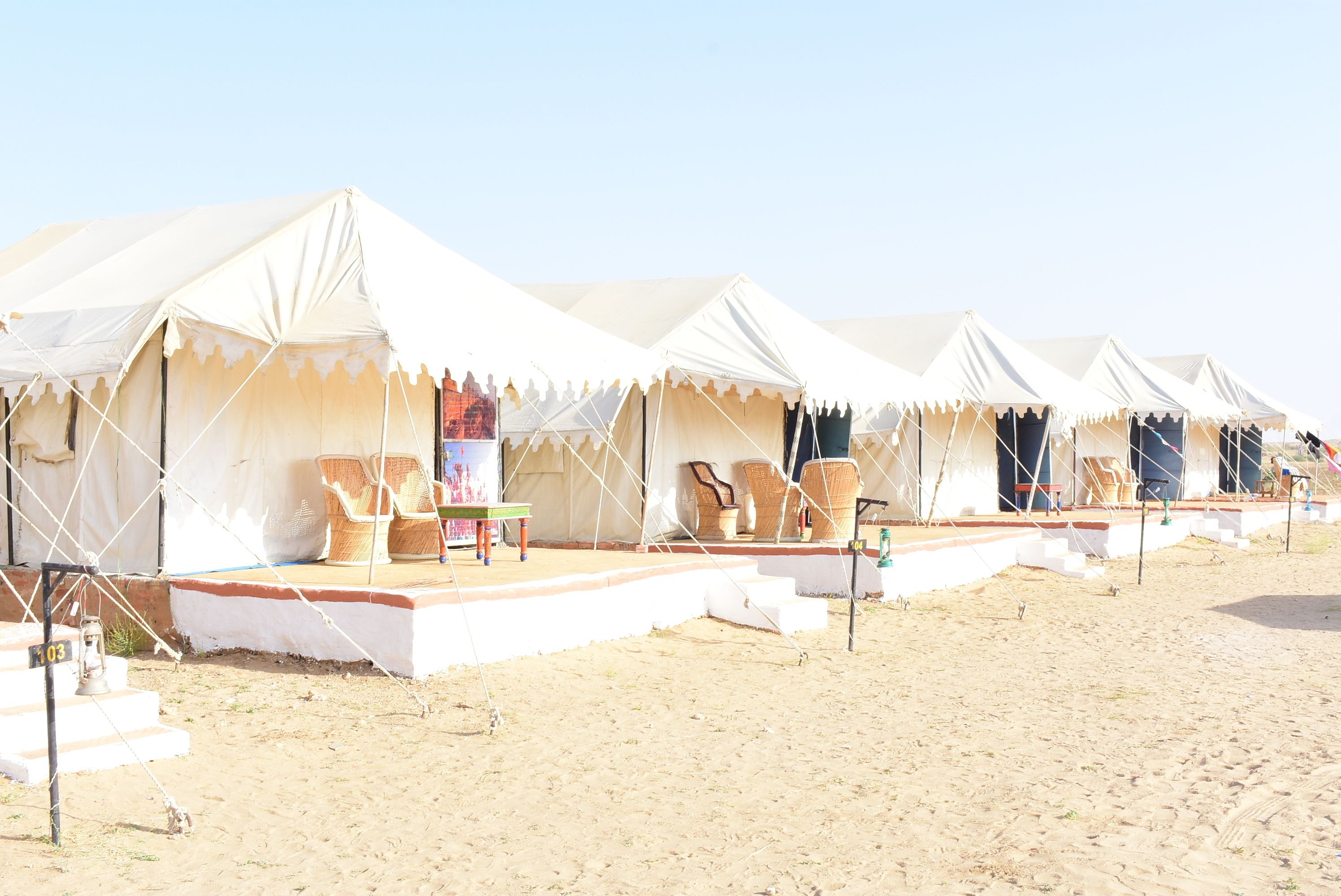 A row of white tents with steps leading up to a stage area, surrounded by beach chairs and tables.