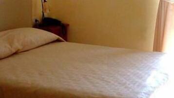 A bed with a beige bedspread in a room with a plain wall and a window with curtains.