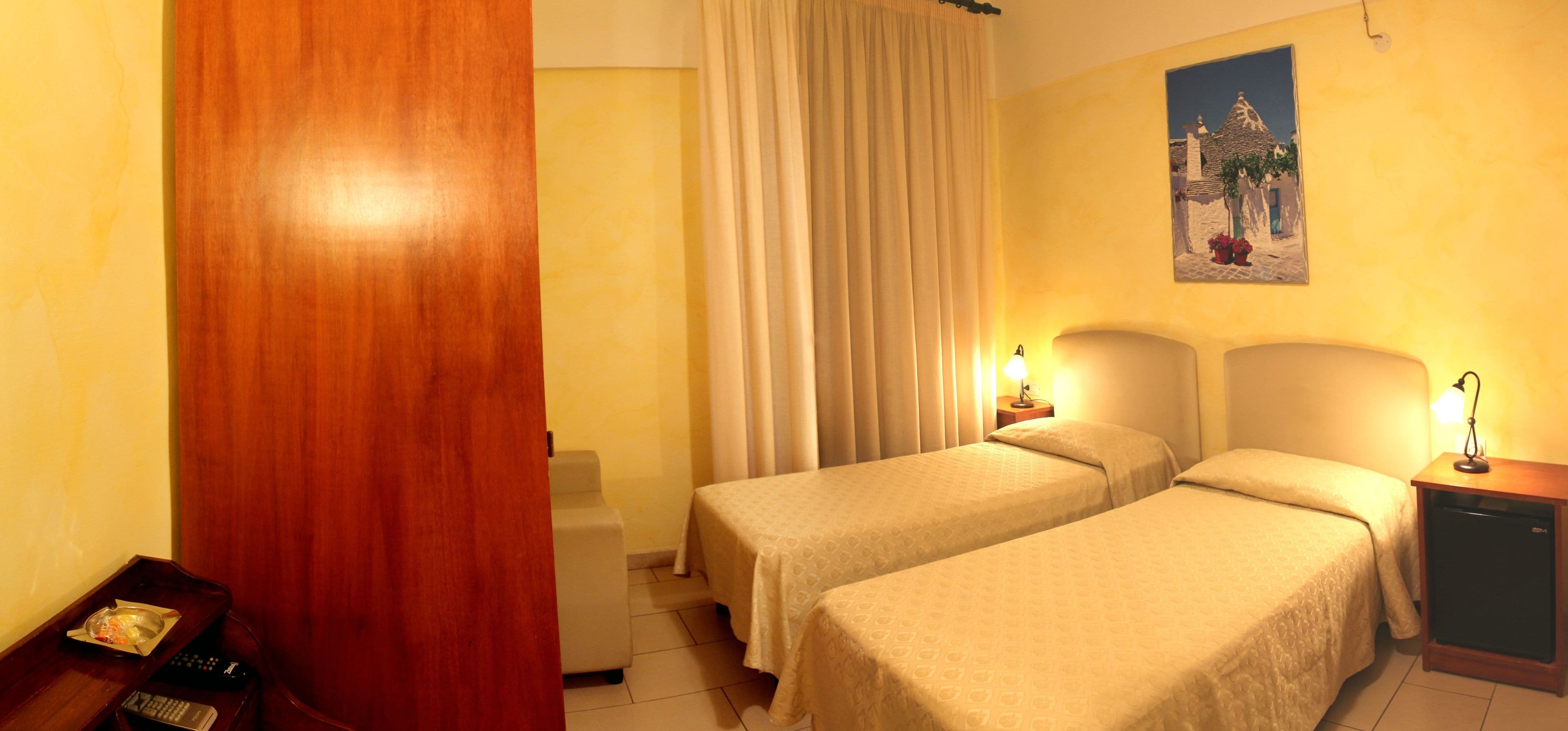 Une chambre d’hôtel avec deux lits simples, une porte en bois, un petit bureau avec une lampe et un tableau au mur.