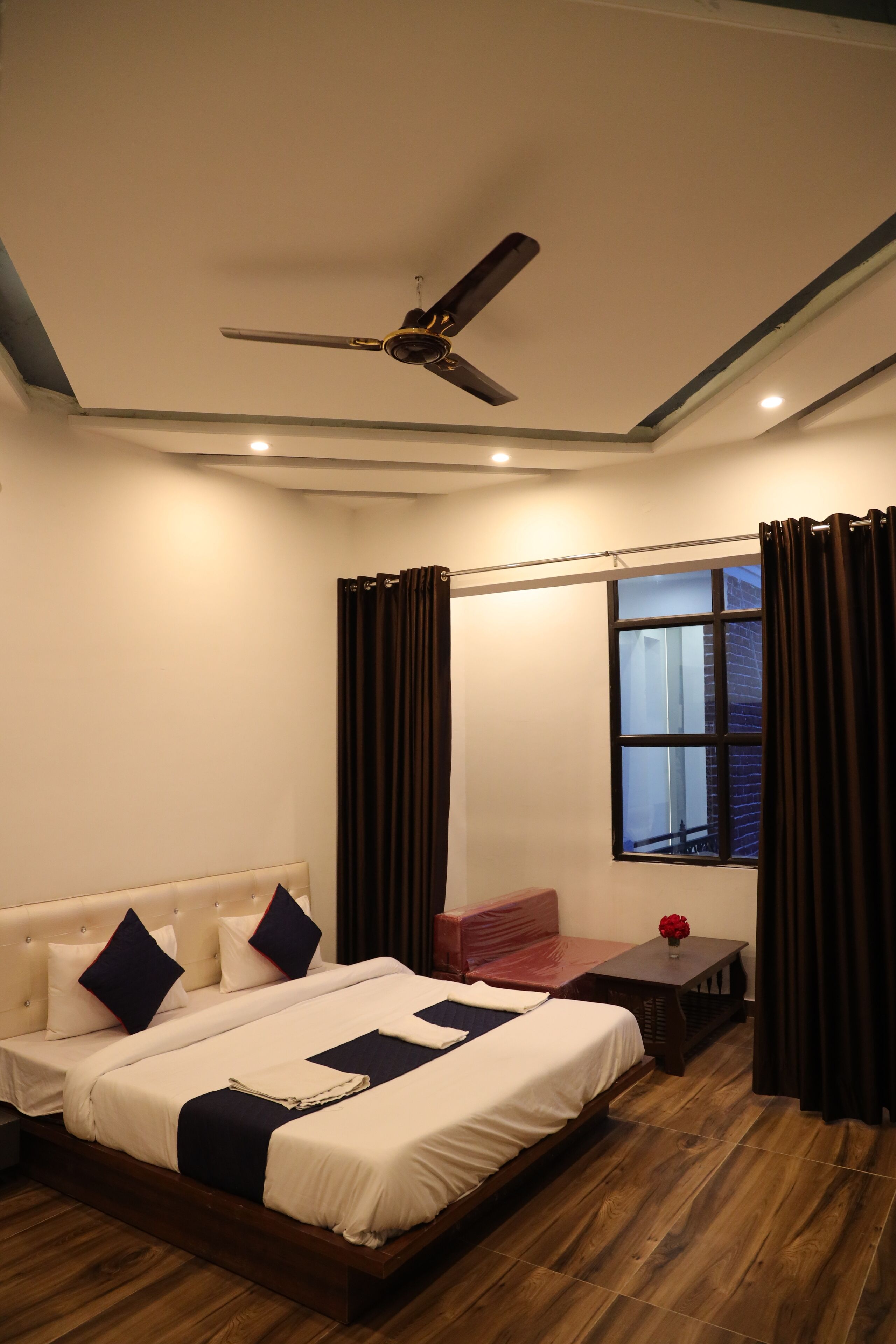 A bedroom with a bed, a ceiling fan, a window with curtains, and a small table with a vase.
