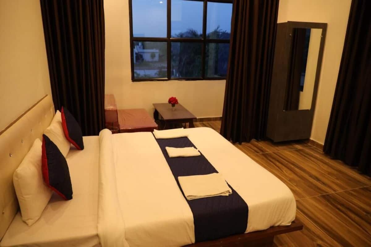 A hotel room with a bed, a small table with a vase, a window, and a door.