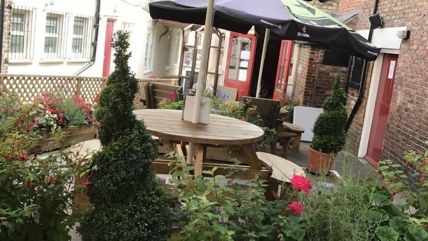An outdoor patio area with a round wooden table and chairs, surrounded by potted plants and a large umbrella providing shade.