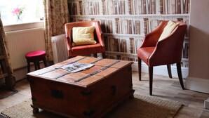 A room with a vintage suitcase, two leather armchairs, a red stool, and a bookshelf filled with books.