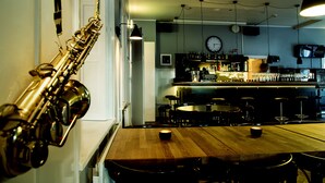 Un coin bar avec une table en bois, des chaises et un saxophone. On y trouve également des lampes suspendues, une horloge et un téléviseur accroché au mur.