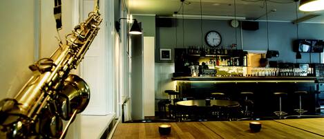 A bar area with a wooden table, chairs, and a saxophone. There are hanging lights, a clock, and a television on the wall.