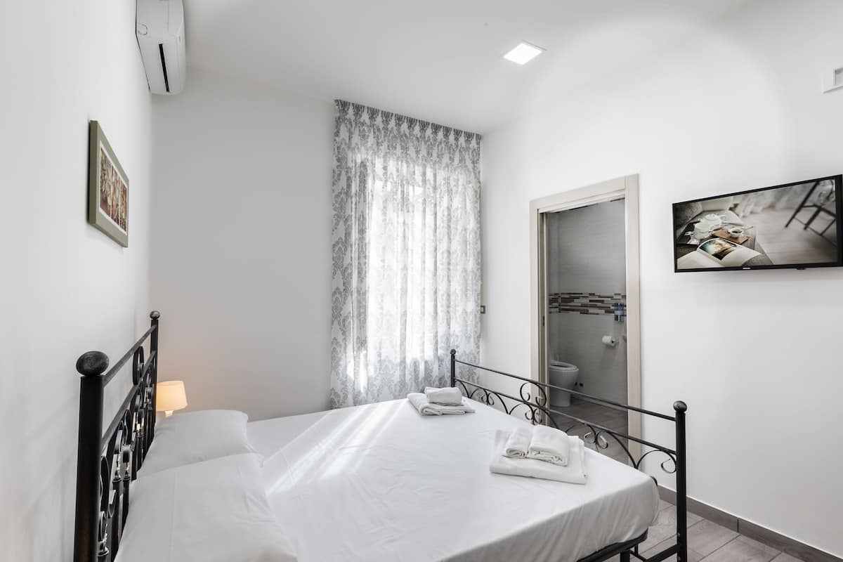 A bedroom with a bed, a bedside table with a lamp, a window with curtains, a painting on the wall, and a bathroom door.
