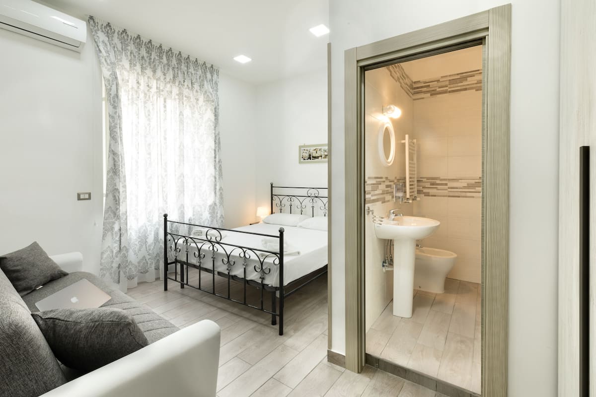 A bedroom with a bed, a bedside table, a chair, and a bathroom with a sink and toilet.