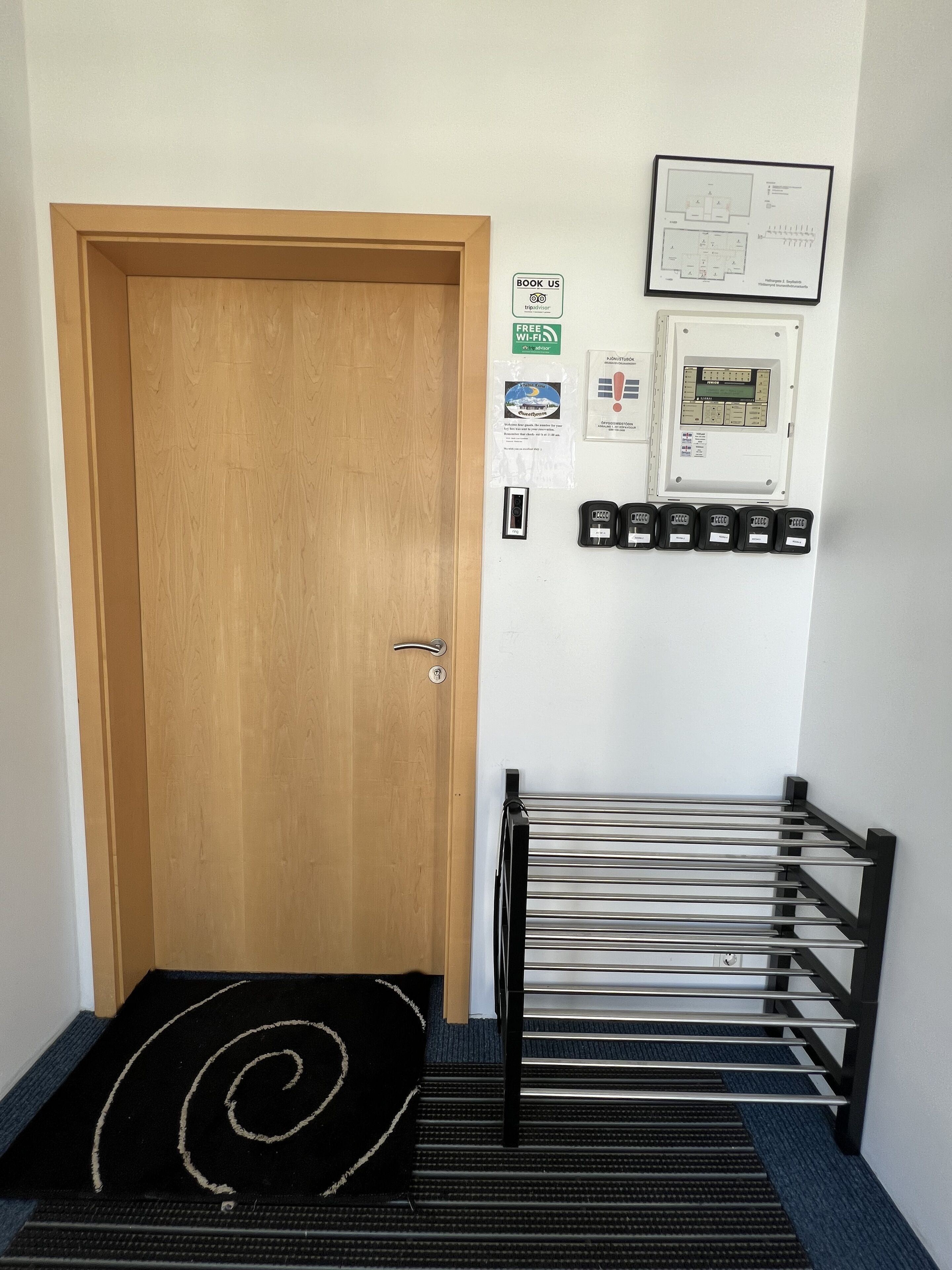 Een hotelingang met een houten deur, een zwarte mat met een spiraalpatroon, een metalen leuning en een elektrische kast aan de muur.