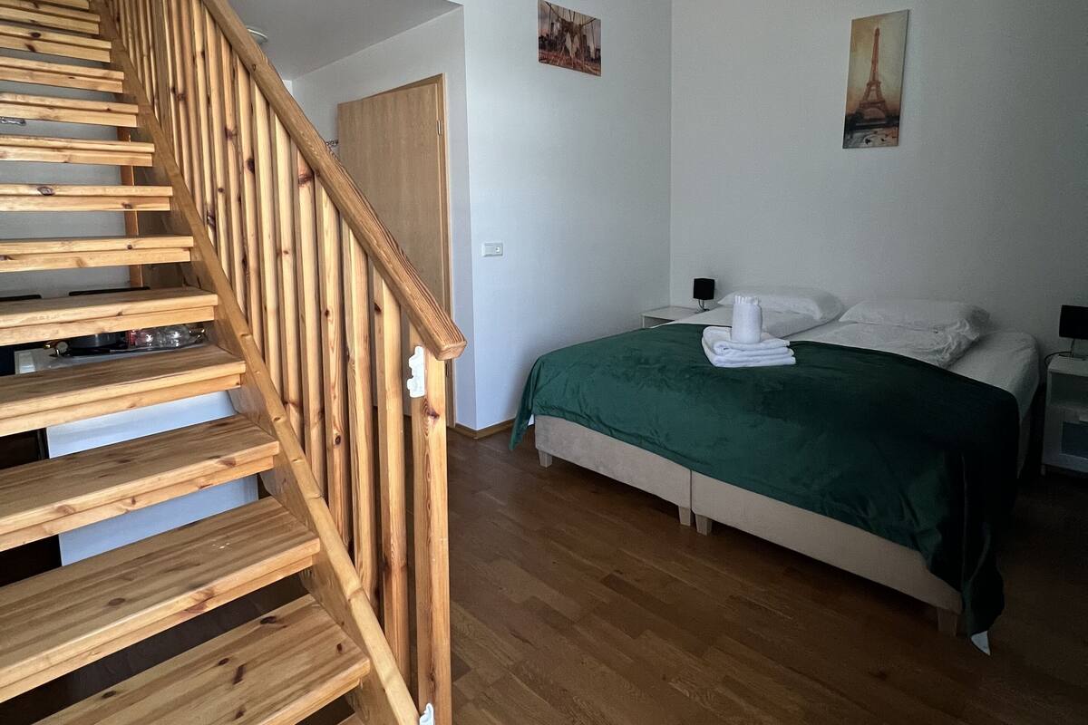 Une chambre avec un escalier en bois, un lit recouvert d’une couverture verte et deux tableaux accrochés au mur.