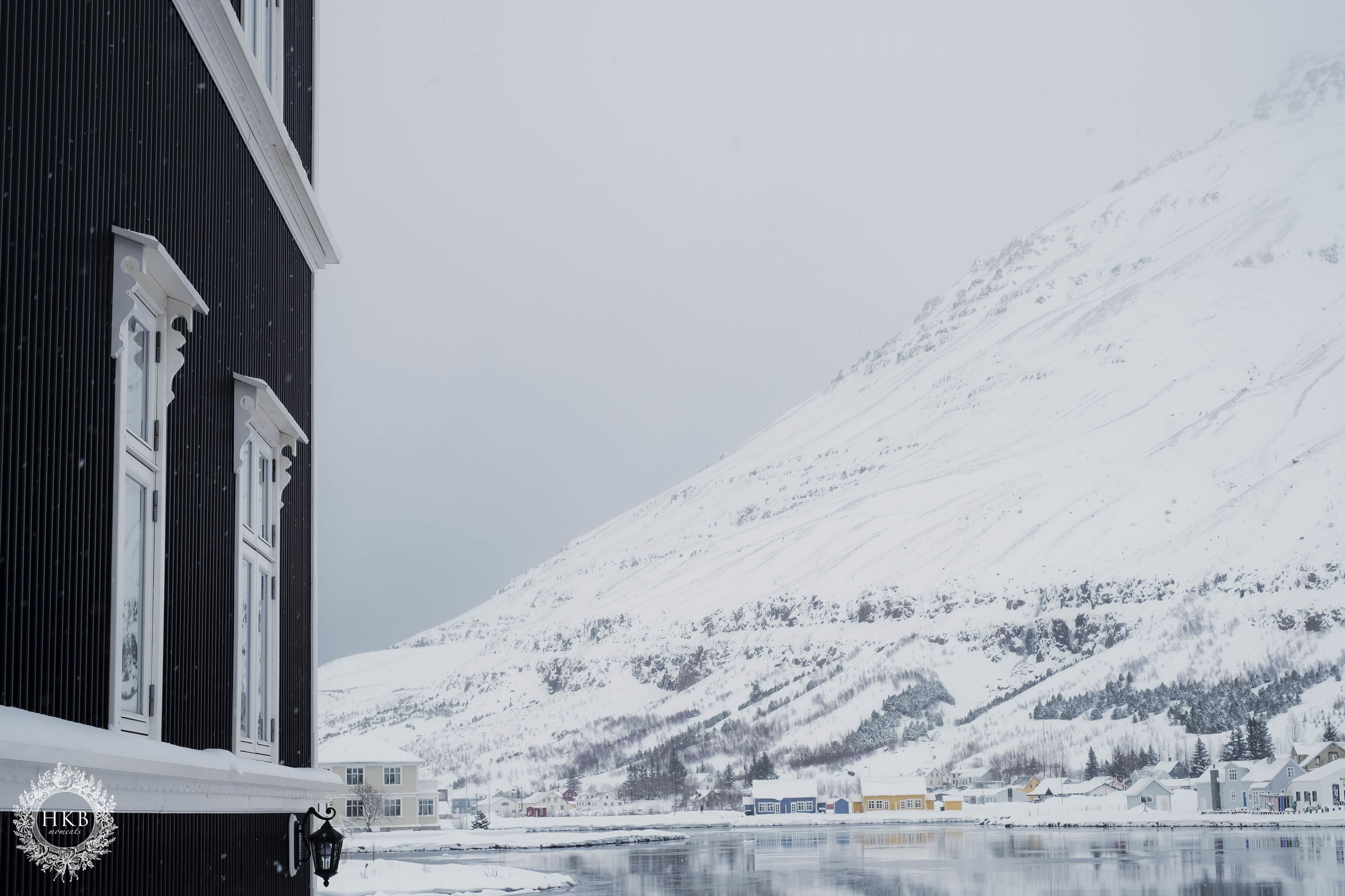 A building with a dark facade and white trim, a snow-covered mountain in the background, and a calm body of water in the foreground.
