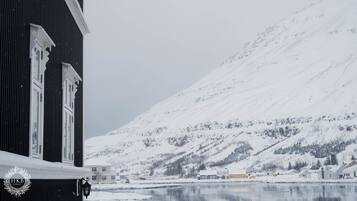 A building with a dark facade and white trim, a snow-covered mountain in the background, and a calm body of water in the foreground.