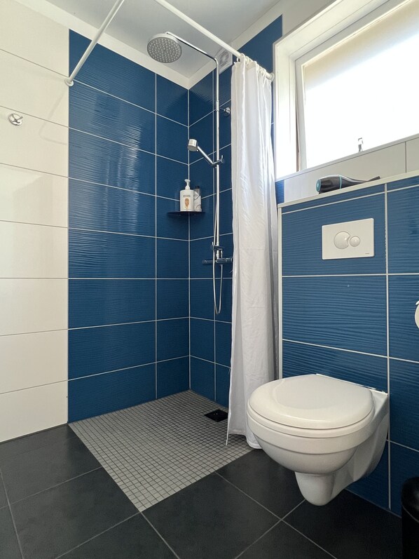 Un baño moderno con paredes de azulejos azules, un inodoro blanco, una ducha con cortina y un pequeño estante con artículos de aseo.