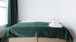 Un lit bien fait, avec une couvre-lit verte et des oreillers blancs, une lampe de chevet blanche et une fenêtre ornée d’un rideau vert.