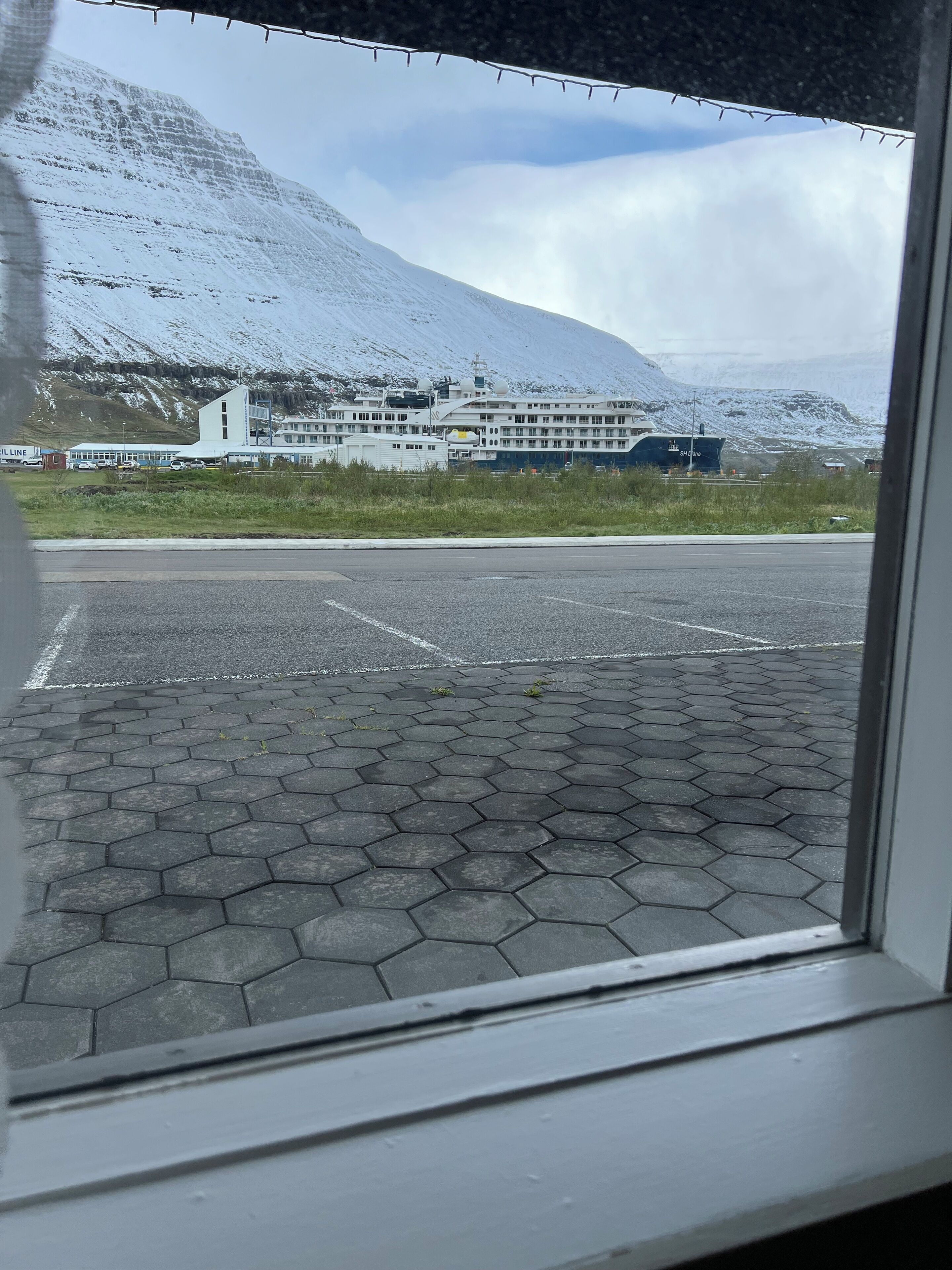 A view of a building with a large ship docked in front, a parking lot with hexagonal paving, and a mountainous backdrop.