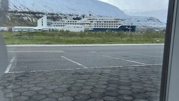A view of a building with a large ship docked in front, a parking lot with hexagonal paving, and a mountainous backdrop.