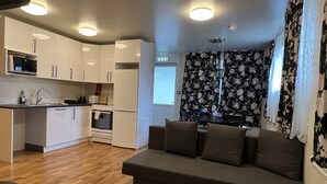Une cuisine moderne avec des meubles blancs, un four à micro-ondes, une cuisinière et un évier. Le salon est équipé d’un canapé gris agrémenté de coussins.