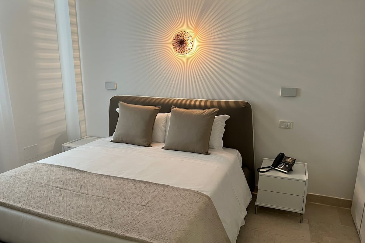 A neatly made bed with a headboard, a bedside table with a phone, and a wall-mounted light fixture.
