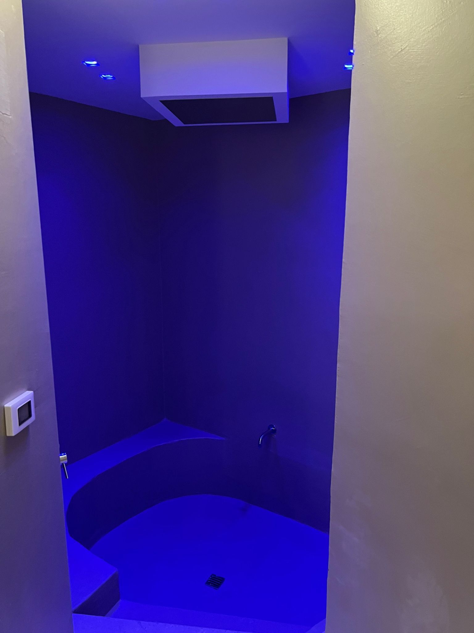 Un baño moderno con zona de ducha iluminada en azul y un respiradero blanco en el techo.