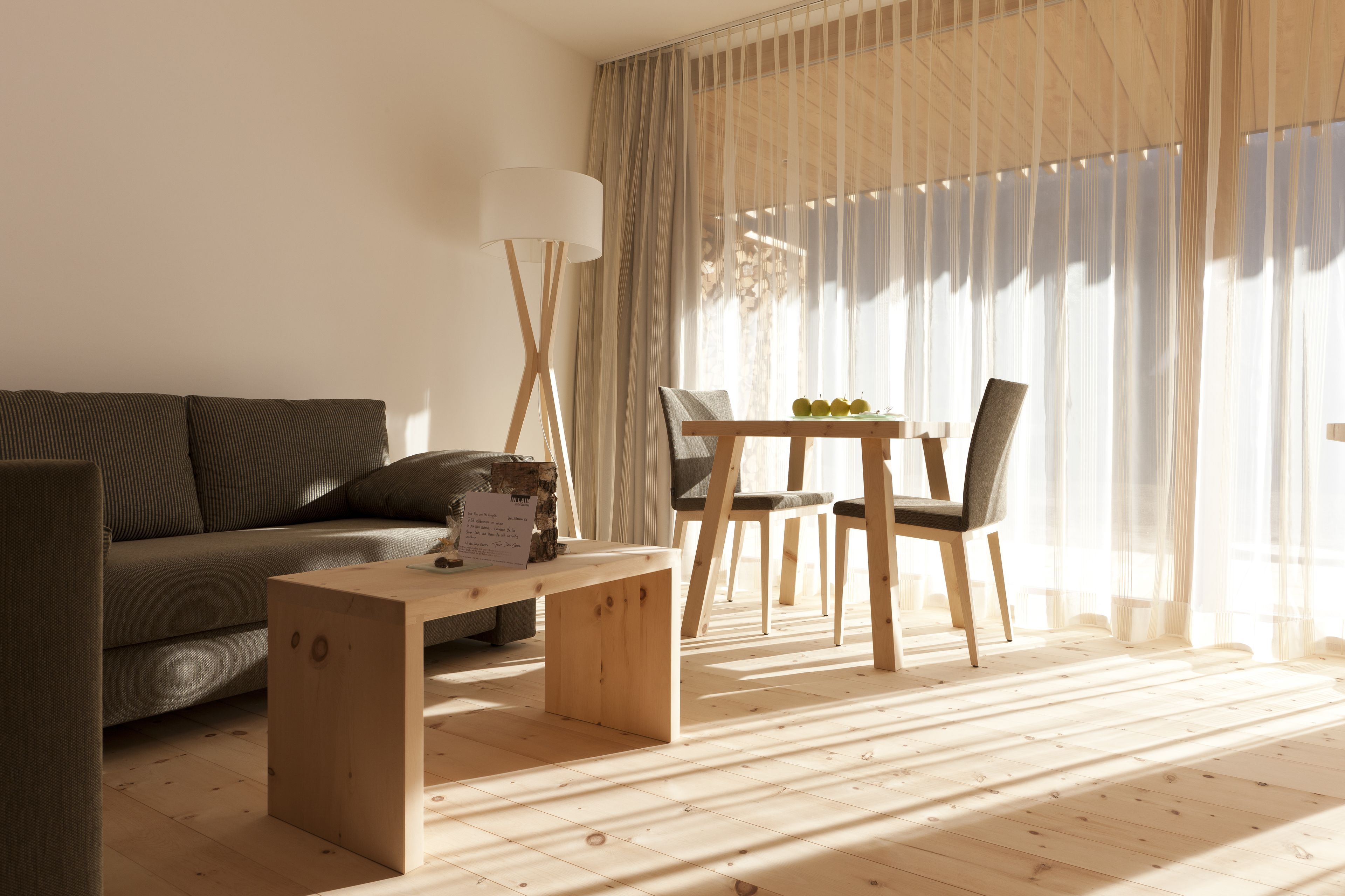 Ein modernes Wohnzimmer mit einer Couch, einem Holztisch, einem Esstisch mit Stühlen und transparenten Vorhängen, die natürliches Licht hereinlassen.