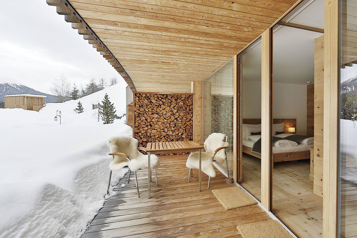 Une terrasse en bois avec une porte vitrée donnant sur une chambre à coucher équipée d’un lit et d’une table de chevet.
