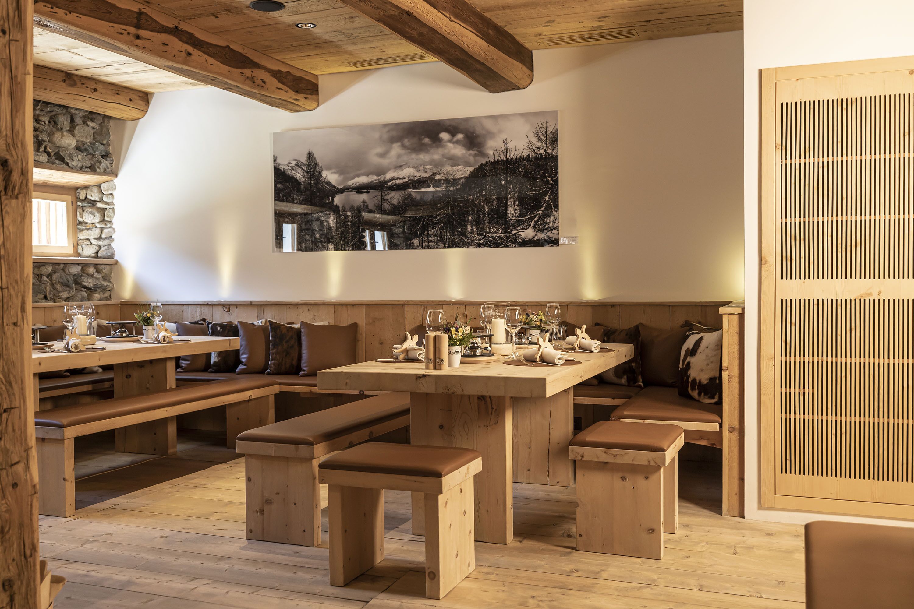 Un interior rústico de madera con una mesa de comedor para cuatro personas, bancos y un cuadro grande con un paisaje en la pared.