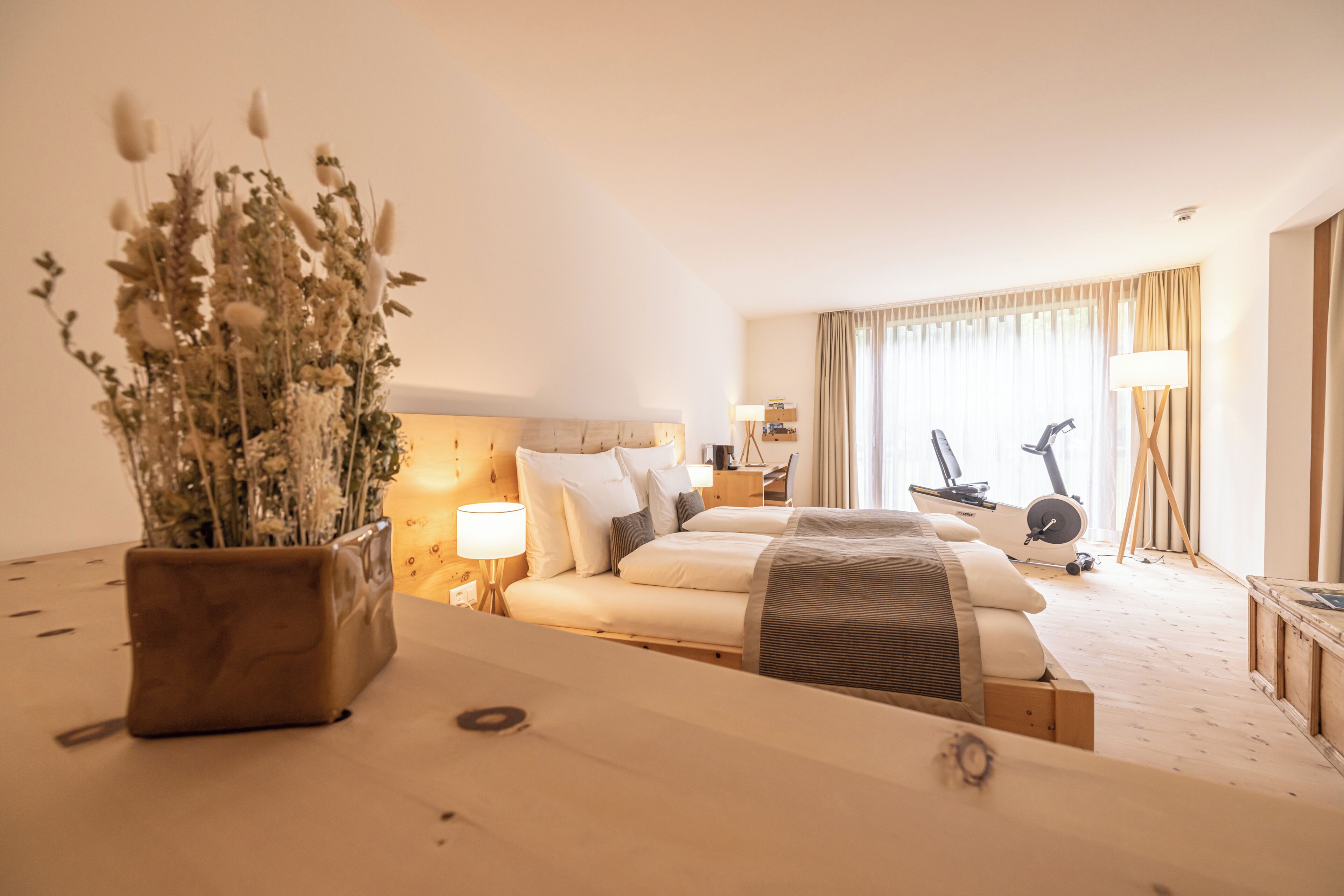 Habitación de hotel con cama, mesita de noche, lámpara, cinta de correr y planta en maceta.