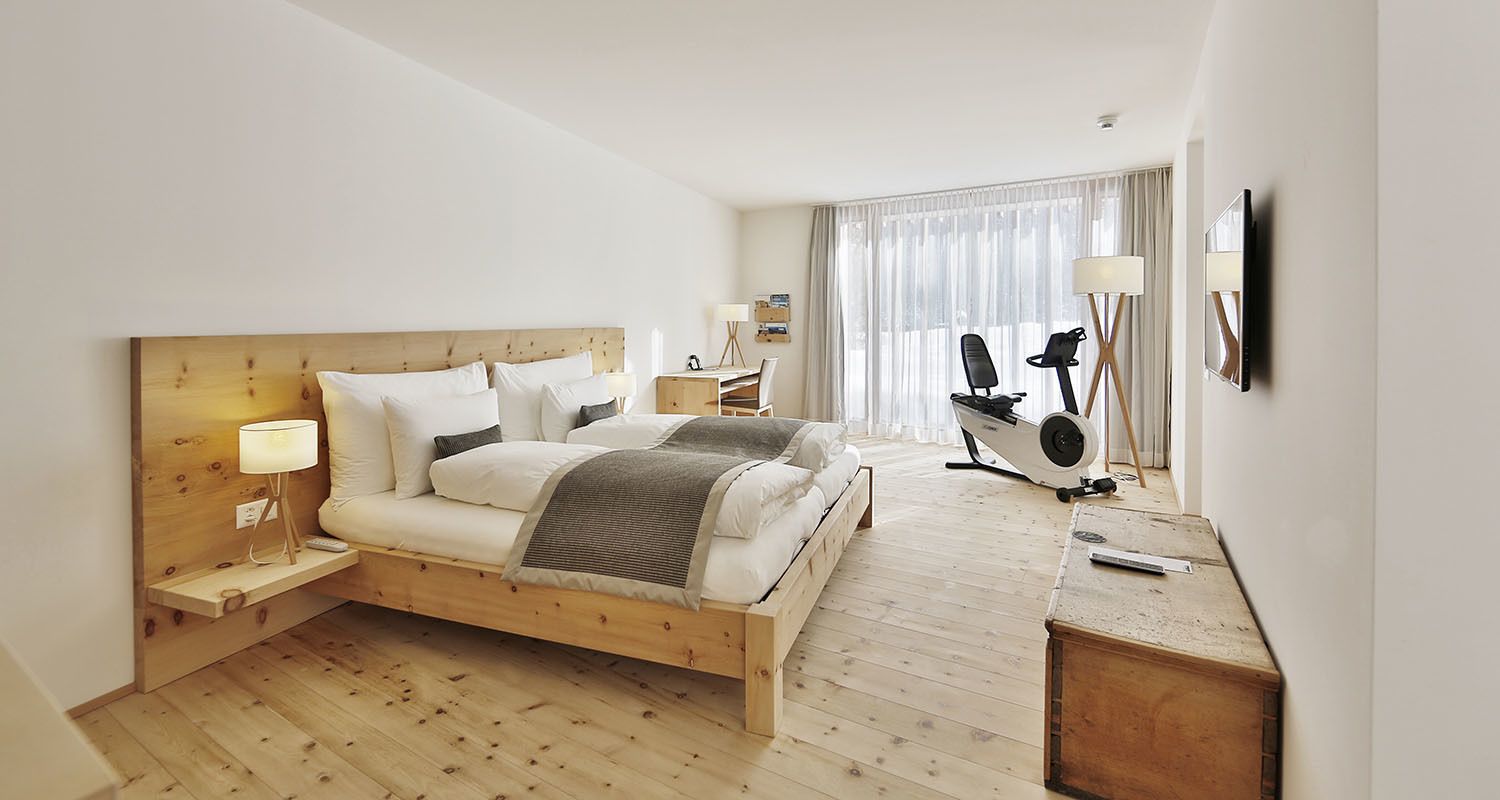 Habitación de hotel moderna con una cama de madera, un escritorio con silla, una cinta de correr y un televisor montado en la pared.
