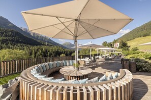 Une terrasse circulaire en bois, dotée d’un espace salon intégré, d’un grand parasol et offrant une vue sur les montagnes.