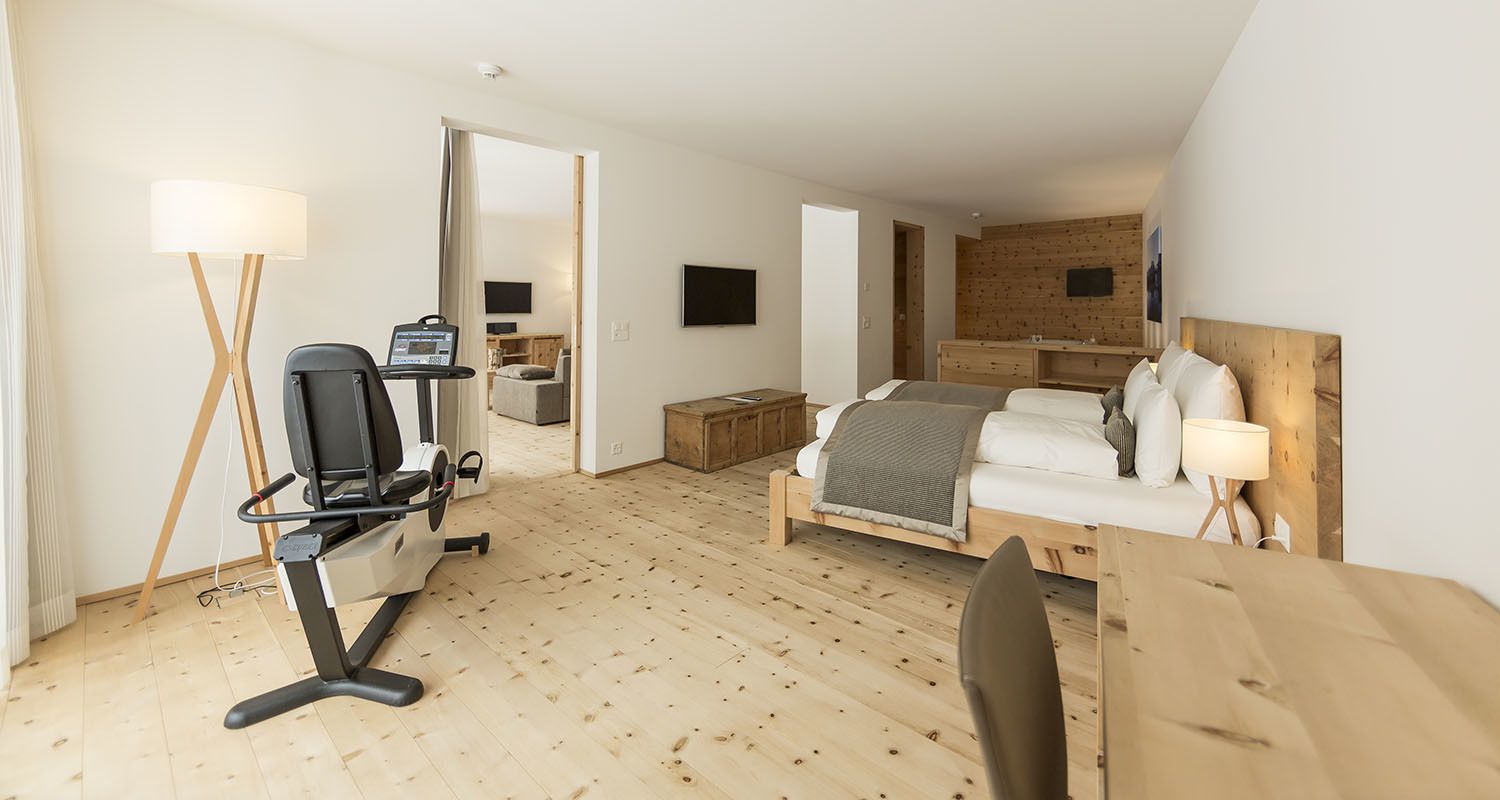 Una habitación de hotel moderna con suelo de madera, cama con ropa blanca, un escritorio con computadora, una silla y una lámpara.
