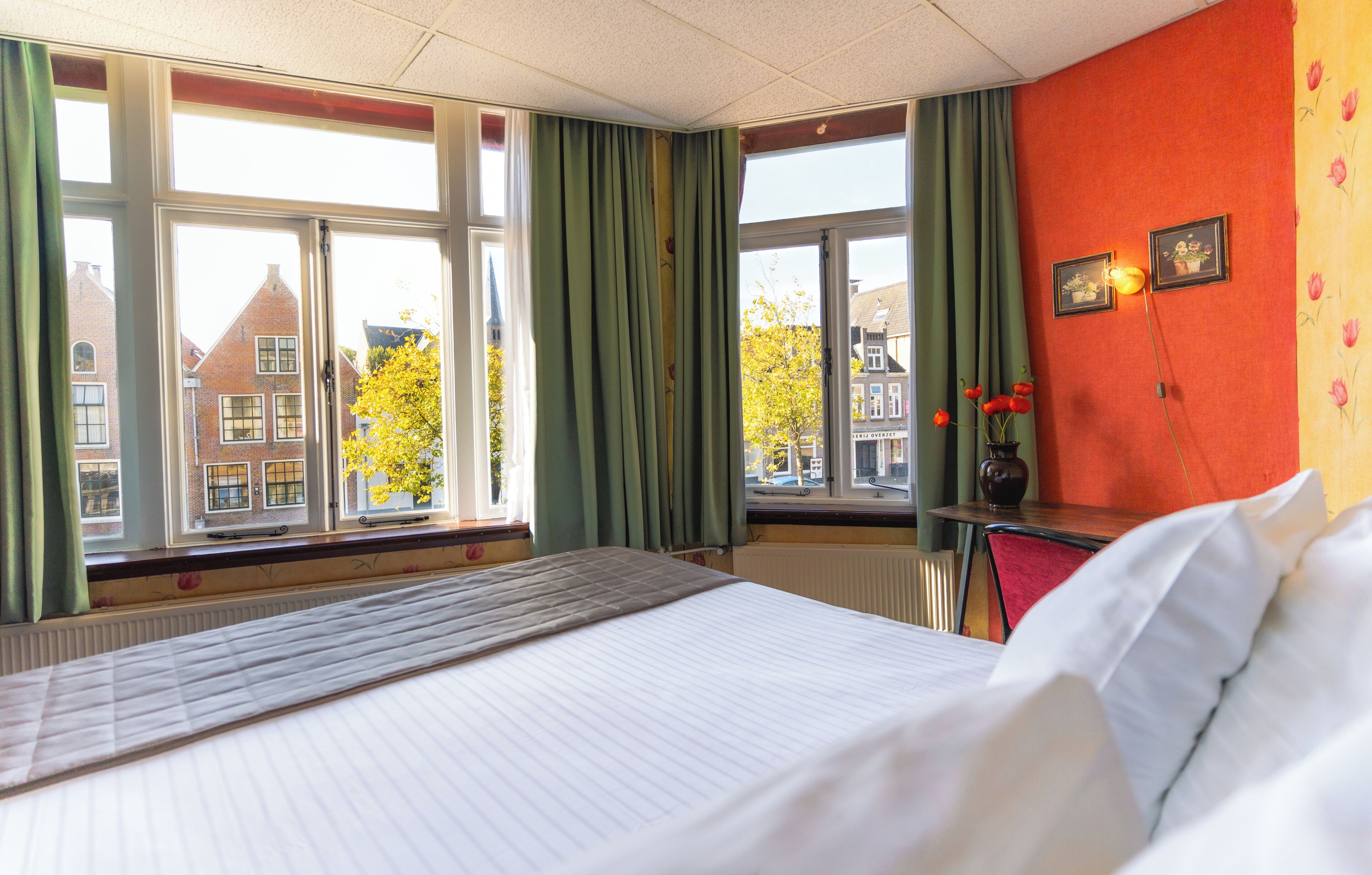 A hotel room with a bed, a window with curtains, a radiator, and a view of buildings outside.