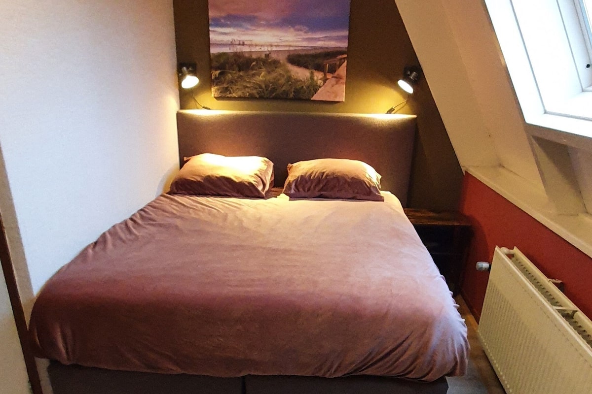 A bedroom with a bed, a nightstand, a wall-mounted lamp, a radiator, and a window.