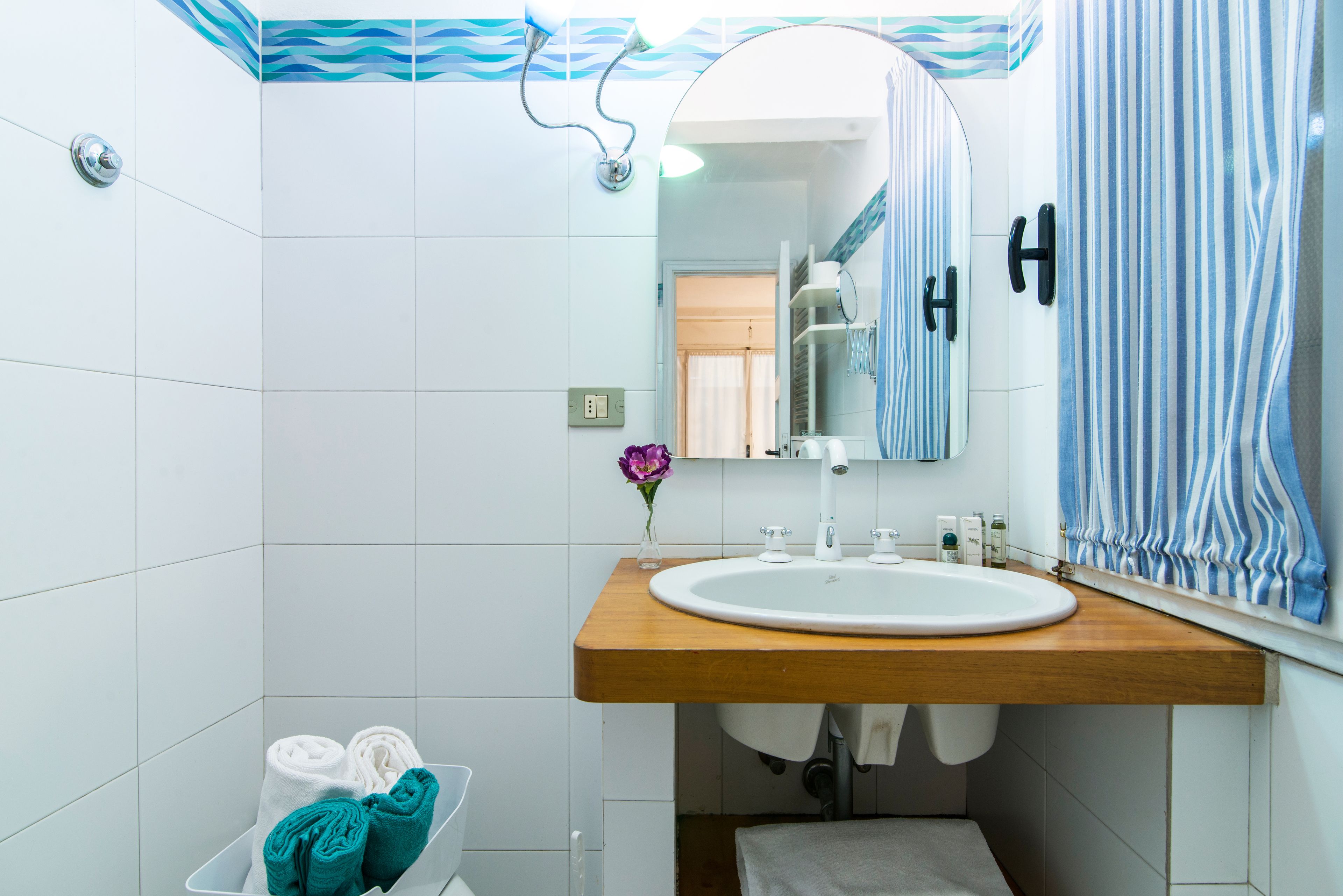 A bathroom with a white sink, wooden countertop, and a mirror above it. There is a towel rack with towels and a small vase with a flower.