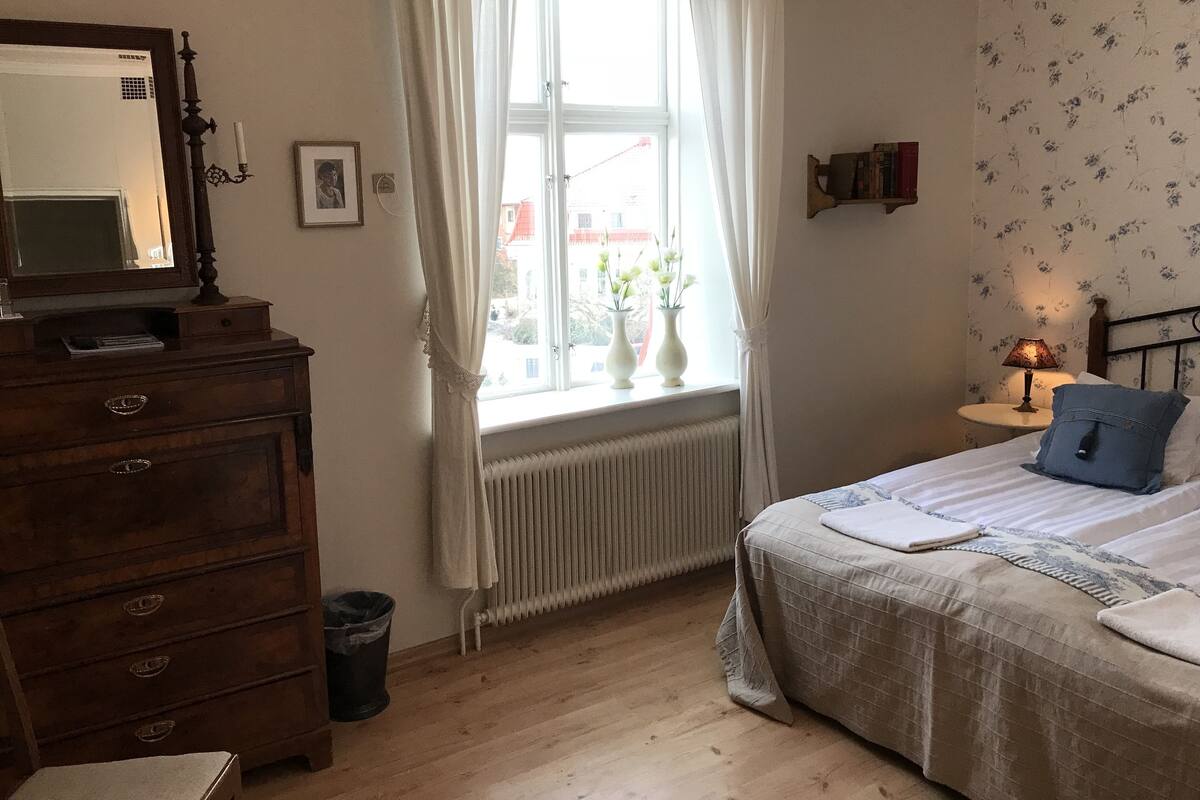 Um quarto com cômoda de madeira, cama com travesseiro azul, janela com cortinas brancas, radiador e uma mesa pequena com abajur.