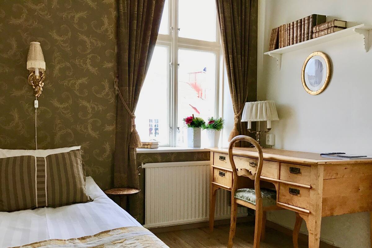 Quarto com cama, escrivaninha de madeira, cadeira, radiador, janela com cortinas e estante com livros.