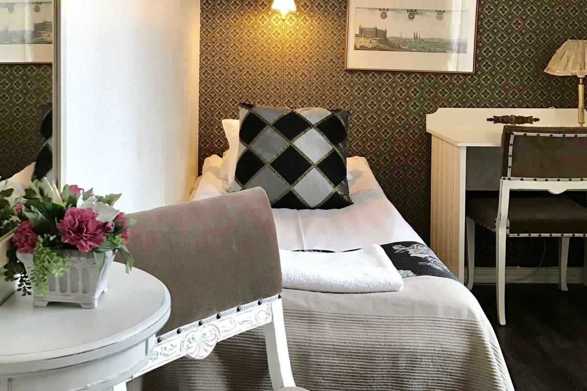 Quarto de hotel bem arrumado com uma cama, uma mesa pequena com abajur, uma cadeira e um vaso decorativo com flores.