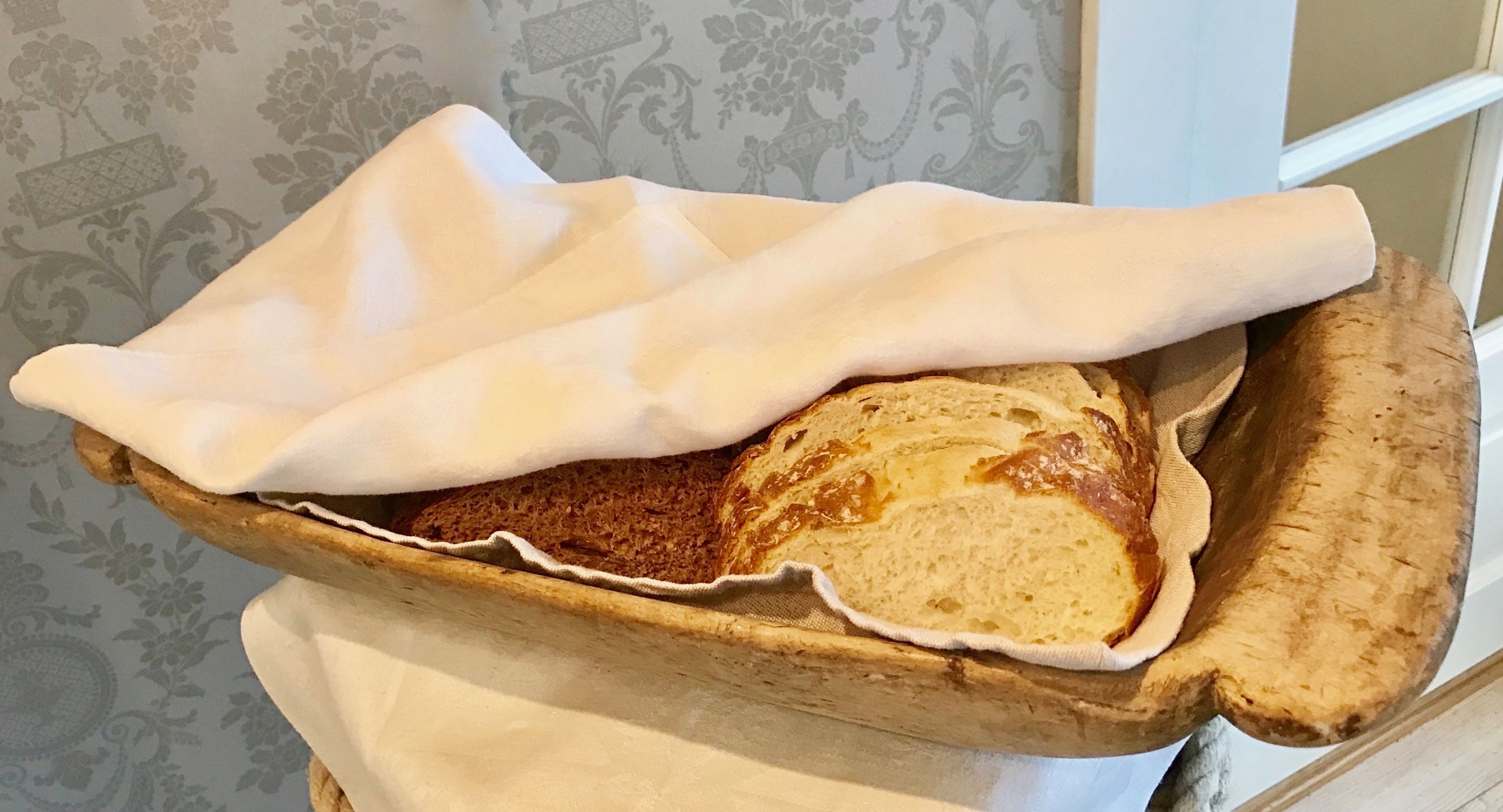 A wooden bread basket with a cloth cover, placed on a table with a patterned wallpaper background.