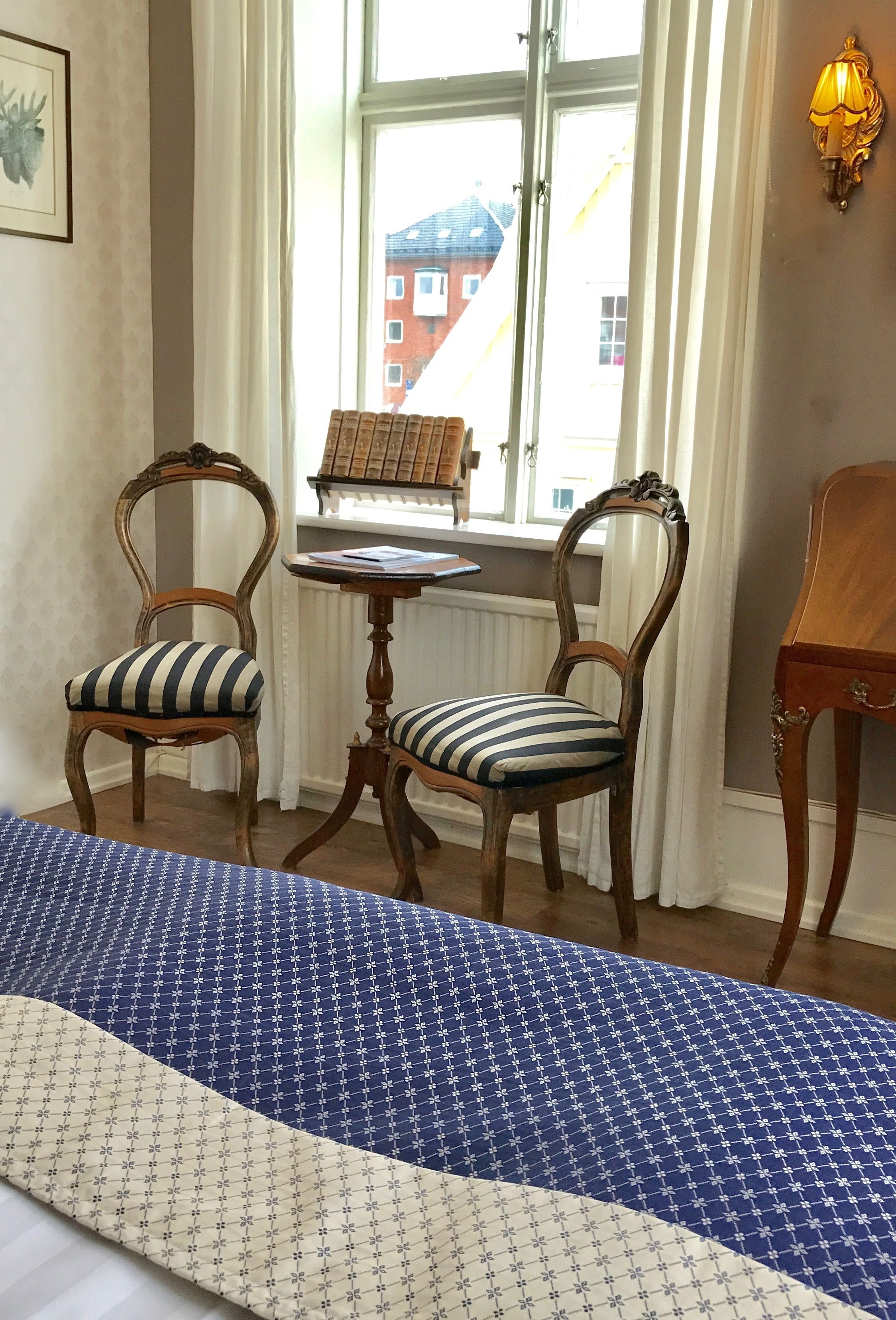 Una habitación con dos sillas de madera con cojines a rayas, una mesita redonda pequeña y una cama con edredón azul y blanco.