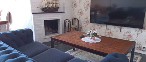 Una sala de estar con un amplio sofá azul, una mesa de centro de madera con un jarrón floral, una chimenea decorada con un modelo de barco y un televisor de pantalla plana montado en la pared.