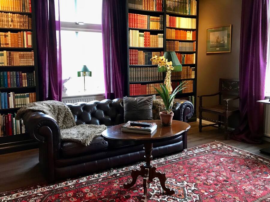 A cozy living room with a leather sofa, a coffee table, a bookshelf, and a large window with curtains.