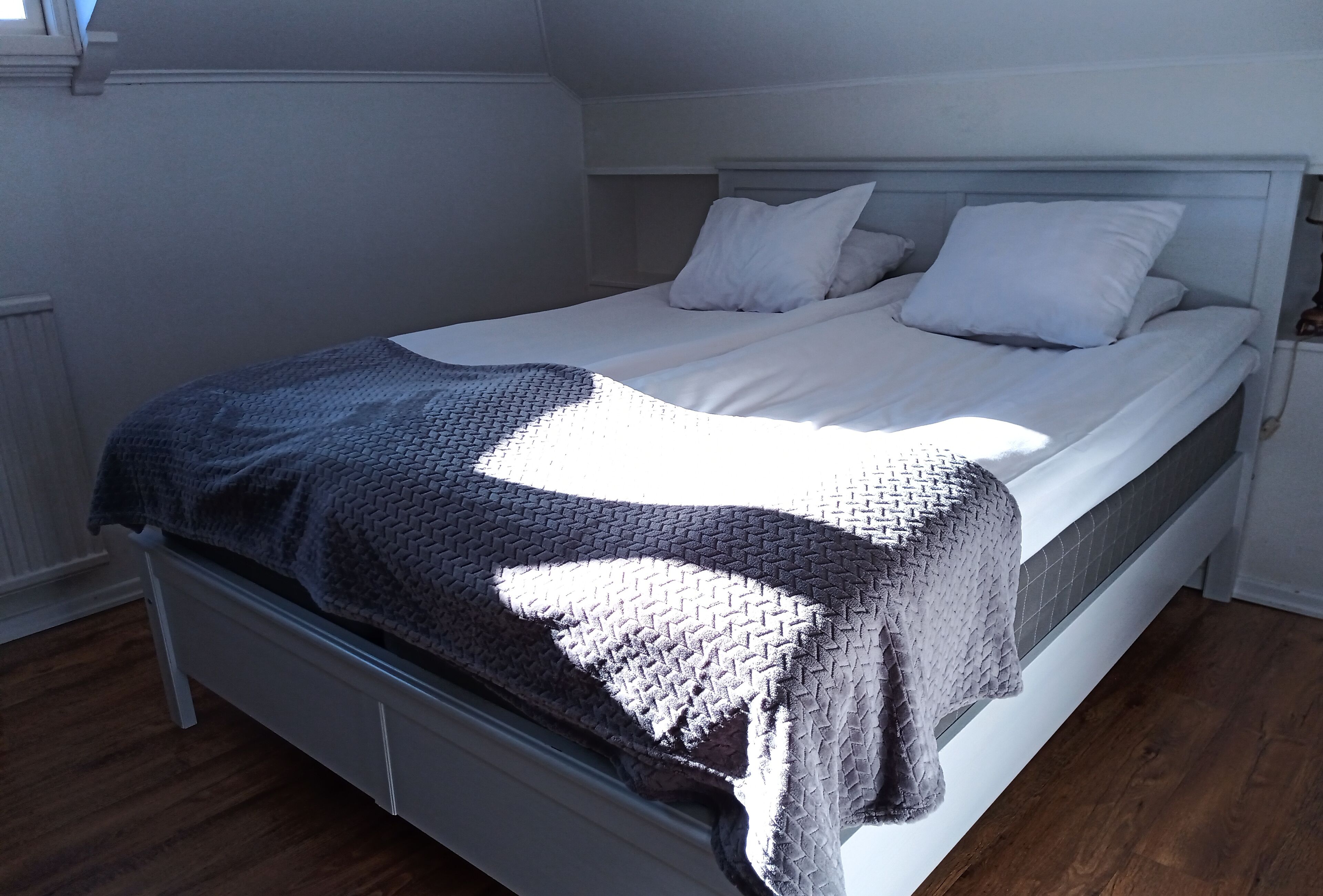 A neatly made bed with a gray and white patterned blanket, white pillows, and a white headboard in a room with wooden flooring and white walls.