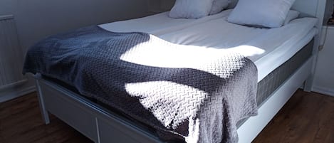 A neatly made bed with a gray and white patterned blanket, white pillows, and a white headboard in a room with wooden flooring and white walls.